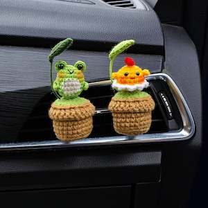 May include: Two handmade crochet car air vent decorations. One is a green frog with a white belly and the other is a yellow chick with a red comb. Both are in brown pots with green leaves.