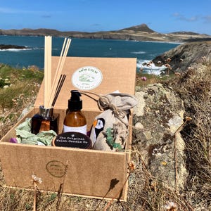 Könnte beinhalten: Eine braune Pappschachtel gefüllt mit Pflegeprodukten, darunter ein Diffusor, eine Flasche mit Pumpe und ein kleiner Beutel. Die Schachtel ist mit "Mor Calm" und "The Original Welsh Candle Co." beschriftet. Der Hintergrund zeigt eine Küstenlandschaft mit blauem Wasser und grünen Hügeln.