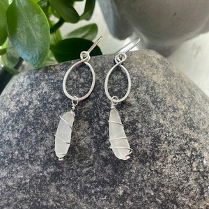 May include: A pair of silver earrings with a white sea glass charm hanging from each. The earrings are made of wire and have a simple, elegant design.