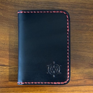 以下が含まれることがあります： 赤いステッチが縁にある黒い革の財布。財布の前面には、エンボス加工された航海用ホイールのエンブレムが付いています。長方形の財布は閉じているように見えます。