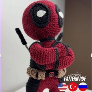 May include: A handmade crochet Deadpool figure in a classic red and black costume. The figure has white eyes, a utility belt, and a black sword. The image also includes the text "crochet PATTERN PDF" and flags.