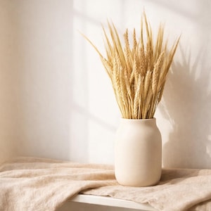 Può includere: Un vaso di ceramica beige pieno di spighe di grano essiccate. Il vaso è appoggiato su un panno di lino dai toni neutri, con un muro bianco sullo sfondo. La scena è immersa in una luce naturale soffusa, creando un'estetica calda.