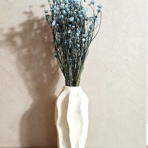 May include: A white, textured vase with a wavy design holds a bundle of dried blue flowers. The vase has a matte finish and a unique, organic shape. The flowers have delicate, small blooms and slender stems. The arrangement is set against a neutral background.