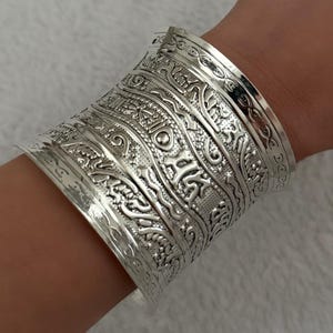 Könnte beinhalten: Ein breites silberfarbenes Manschettenarmband mit komplexen Prägemustern auf der Oberfläche. Das Design besteht aus mehreren horizontalen Bändern mit unterschiedlichen Texturen, darunter geometrische Formen und florale Motive.