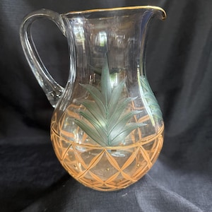 May include: Clear glass pitcher with a handle and a gold rim. The pitcher features a pineapple design with green leaves and a diamond pattern in shades of orange and gold. The pitcher is designed for serving beverages.