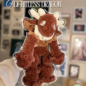 May include: A brown plush dragon toy with cream-colored horns and a ruffled collar, cuffs, and leg accents. The text OZZI THE FLIGHTLESS DRAGON is displayed above the creature, which features a soft, velvet-like texture and white eyes.