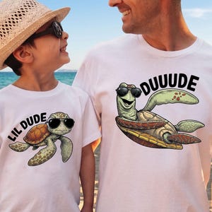 May include: White t-shirts with matching turtle graphics. One shirt says "Lil Dude" and the other says "Duuude". The turtle illustrations wear sunglasses. The shirts are worn by a child and an adult on a beach.