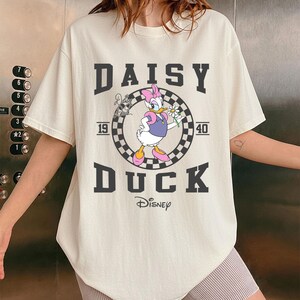 May include: Cream-coloured t-shirt with a graphic of Daisy Duck holding a flower, set within a chequered circle. The text "DAISY DUCK" and "1940" are printed above and below the graphic, with the Disney logo at the bottom.
