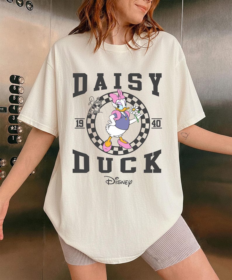 May include: Cream-coloured t-shirt with a graphic of Daisy Duck holding a flower, set within a chequered circle. The text "DAISY DUCK" and "1940" are printed above and below the graphic, with the Disney logo at the bottom.