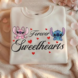 Może przedstawiać: Kremowa bluza z postaciami Angel i Stitch, napisami "Forever Sweethearts" i czerwonymi serduszkami. Projekt jest wyśrodkowany na przodzie bluzy.