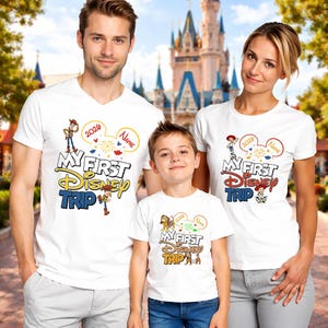 May include: Three people wearing white t-shirts with the text "My First Disney Trip" in a colorful design. The shirts feature Disney characters and the year 2020. The family is posing in front of a castle.