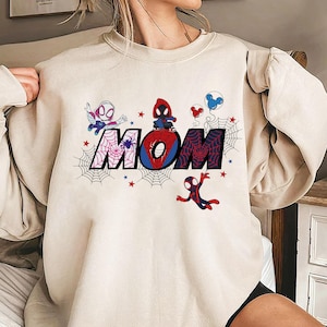 Puede incluir: Sudadera color crema con la palabra "MOM" en un dise&ntilde;o de telara&ntilde;a con personajes de Spider-Man. Las letras est&aacute;n decoradas con telara&ntilde;as, estrellas y personajes de dibujos animados. Un dise&ntilde;o divertido y colorido.