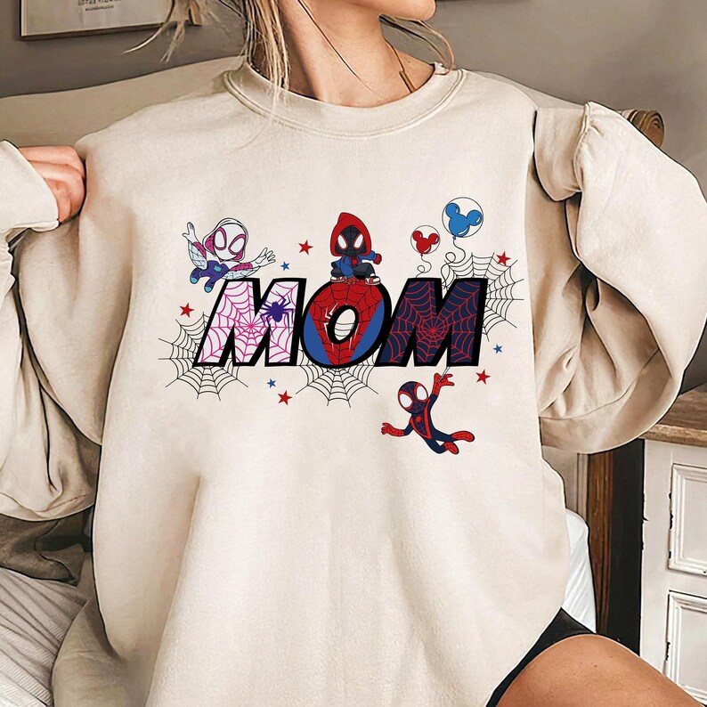 Puede incluir: Sudadera color crema con la palabra "MOM" en un dise&ntilde;o de telara&ntilde;a con personajes de Spider-Man. Las letras est&aacute;n decoradas con telara&ntilde;as, estrellas y personajes de dibujos animados. Un dise&ntilde;o divertido y colorido.