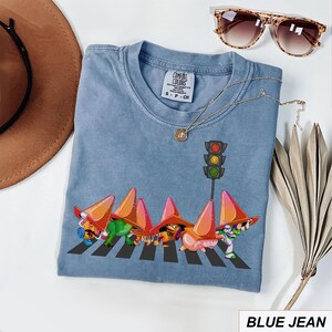 May include: Blue jean Comfort Colors t-shirt featuring a whimsical graphic of cartoon characters crossing a crosswalk under a traffic light. The shirt has a crew neck and short sleeves. A gold necklace and sunglasses are also visible.