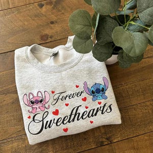 Może przedstawiać: Jasnoszara bluza z postaciami Angel i Stitch, napisami "Forever Sweethearts" i czerwonymi serduszkami. Bluza wykonana jest z miękkiego materiału.