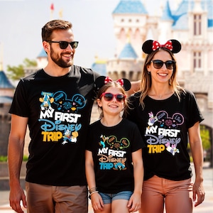 May include: Three people wearing black t-shirts with the text "MY FIRST DISNEY TRIP" in blue, yellow, and white. The shirts feature Disney character graphics and the year 2020. The family is smiling in front of a Disney castle.