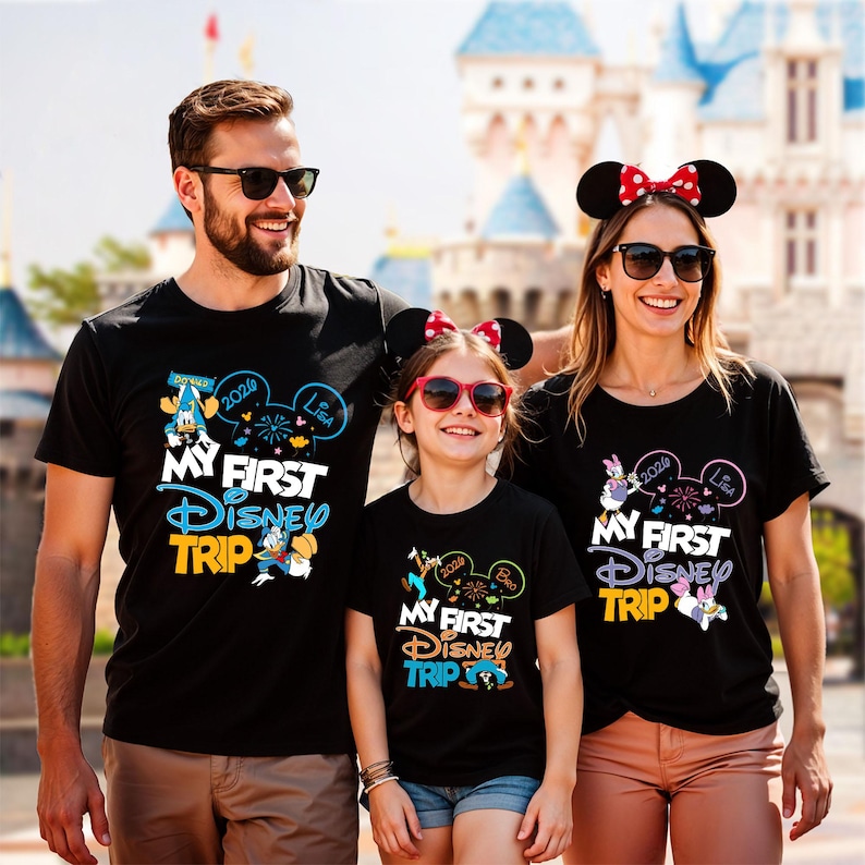 May include: Three people wearing black t-shirts with the text "MY FIRST DISNEY TRIP" in blue, yellow, and white. The shirts feature Disney character graphics and the year 2020. The family is smiling in front of a Disney castle.