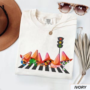 May include: Ivory-colored t-shirt featuring a graphic of cartoon characters crossing a crosswalk under a traffic light. The characters are wearing orange traffic cones. Sunglasses and a necklace are also visible.