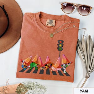 May include: A terracotta-colored Comfort Colors t-shirt featuring a whimsical graphic of cartoon characters crossing a crosswalk under a traffic light. The shirt has a crew neck and short sleeves. A gold necklace and sunglasses are also visible.