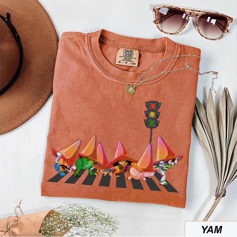 May include: A terracotta-colored Comfort Colors t-shirt featuring a whimsical graphic of cartoon characters crossing a crosswalk under a traffic light. The shirt has a crew neck and short sleeves. A gold necklace and sunglasses are also visible.