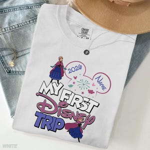 May include: White t-shirt with the text "MY FIRST DISNEY TRIP" in pink and blue, featuring cartoon images of a princess and fireworks. The year "2020" is also displayed. The shirt is a Comfort Colors brand.