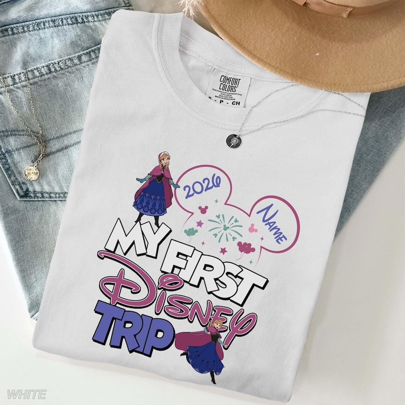 May include: White t-shirt with the text "MY FIRST DISNEY TRIP" in pink and blue, featuring cartoon images of a princess and fireworks. The year "2020" is also displayed. The shirt is a Comfort Colors brand.
