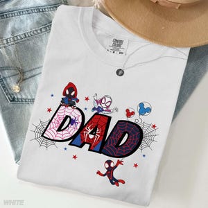 Puede incluir: Camiseta blanca con la palabra "DAD" en estilo c&oacute;mic, con personajes de Spider-Man y dise&ntilde;os de telara&ntilde;as. La camiseta incluye detalles en rojo y azul, estrellas y la etiqueta de la marca "Comfort Colors".