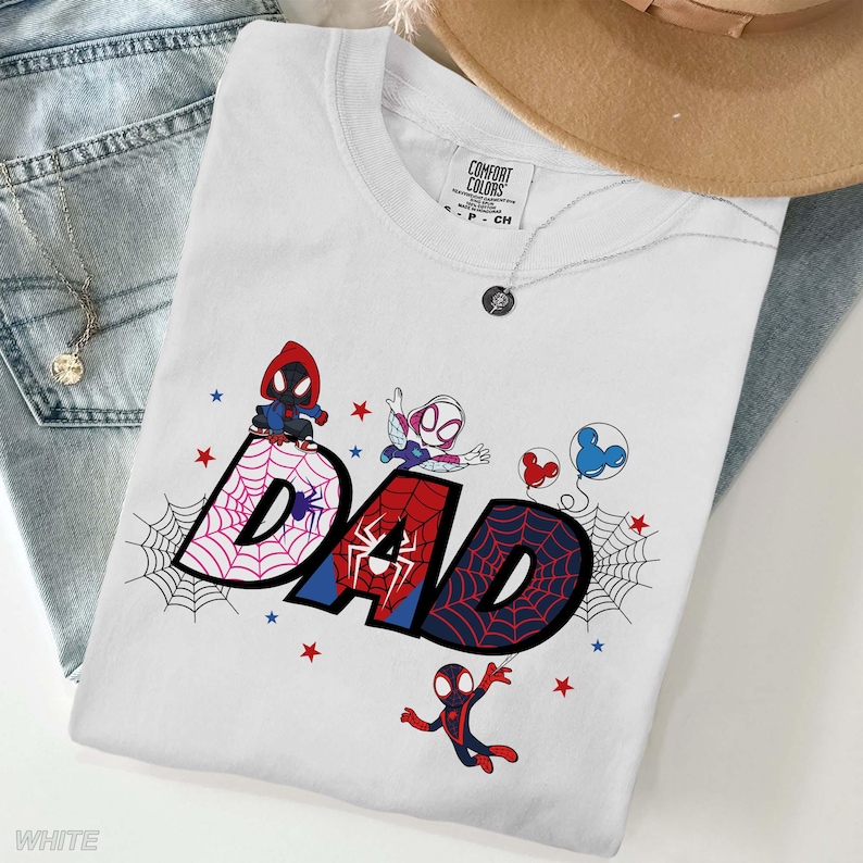 Puede incluir: Camiseta blanca con la palabra "DAD" en estilo c&oacute;mic, con personajes de Spider-Man y dise&ntilde;os de telara&ntilde;as. La camiseta incluye detalles en rojo y azul, estrellas y la etiqueta de la marca "Comfort Colors".