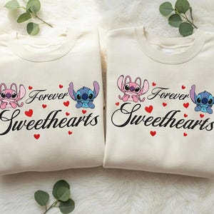 Może przedstawiać: Dwie kremowe bluzy z napisem "Forever Sweethearts" czarnym pismem. Każda koszulka przedstawia postać z kresk&oacute;wki, jedną r&oacute;żową i jedną niebieską, z czerwonymi akcentami w kształcie serca. Motyw romantyczny.