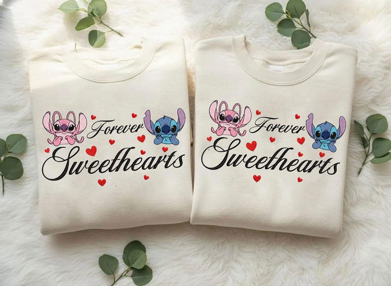 Może przedstawiać: Dwie kremowe bluzy z napisem "Forever Sweethearts" czarnym pismem. Każda koszulka przedstawia postać z kresk&oacute;wki, jedną r&oacute;żową i jedną niebieską, z czerwonymi akcentami w kształcie serca. Motyw romantyczny.