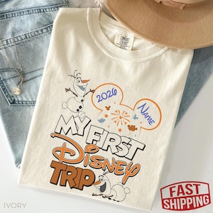 May include: Ivory-colored t-shirt with the text "My First Disney Trip" in orange and white, featuring Olaf the snowman. The design includes a Mickey Mouse ear shape with the year 2026 and space for a name. The shirt is laid on top of blue jeans.