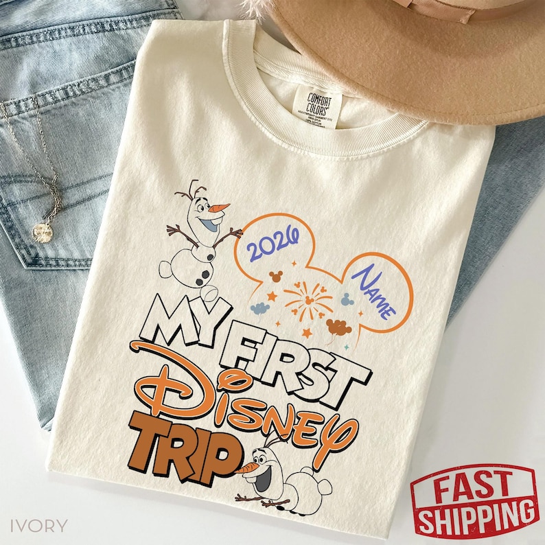 May include: Ivory-colored t-shirt with the text "My First Disney Trip" in orange and white, featuring Olaf the snowman. The design includes a Mickey Mouse ear shape with the year 2026 and space for a name. The shirt is laid on top of blue jeans.