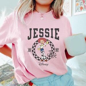 May include: A pale pink sweatshirt with the name "JESSIE" in black, bold lettering. Beneath the name is a circular graphic with a cartoon image of Jessie from Toy Story, the years "19" and "97", and the Disney logo.