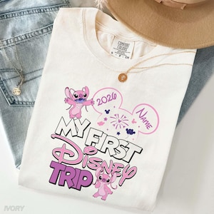 May include: Ivory-colored t-shirt with a Disney theme. The shirt features the text "My First Disney Trip" in a pink and white design, along with the year "2026" and a space for a name. Two pink cartoon characters are also featured.