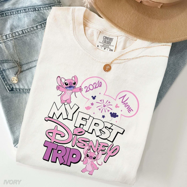 May include: Ivory-colored t-shirt with a Disney theme. The shirt features the text "My First Disney Trip" in a pink and white design, along with the year "2026" and a space for a name. Two pink cartoon characters are also featured.