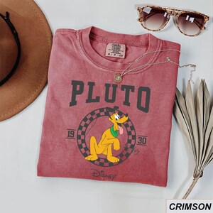 May include: Crimson t-shirt with the word "PLUTO" in dark grey block letters and a cartoon Pluto graphic. The graphic includes the years "19" and "30" and the Disney logo. The shirt is styled with sunglasses and a necklace.