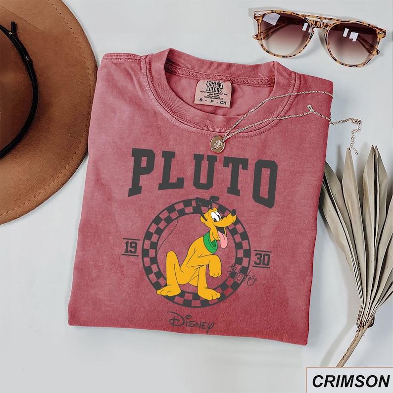 May include: Crimson t-shirt with the word "PLUTO" in dark grey block letters and a cartoon Pluto graphic. The graphic includes the years "19" and "30" and the Disney logo. The shirt is styled with sunglasses and a necklace.