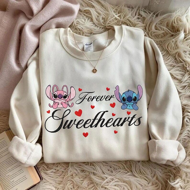 Może przedstawiać: Kremowa bluza z postaciami Angel i Stitch, z napisami "Forever Sweethearts" i czerwonymi sercami. Bluza ma długie rękawy i okrągły dekolt.