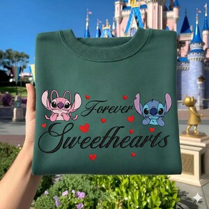 Może przedstawiać: Ciemnozielona bluza z postaciami Stitcha i Angel, napisem "Forever Sweethearts" i czerwonymi serduszkami. Bluza ma okrągły dekolt i długie rękawy. Projekt jest wyśrodkowany z przodu.