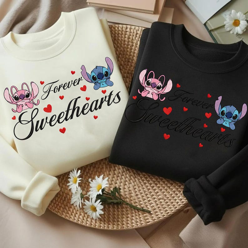 Może przedstawiać: Dwie bluzy z okrągłym dekoltem, jedna kremowa i jedna czarna, z kresk&oacute;wkowym wzorem dw&oacute;ch postaci i napisem "Forever Sweethearts". Projekt zawiera czerwone serca i postacie w kolorze r&oacute;żowym i niebieskim.