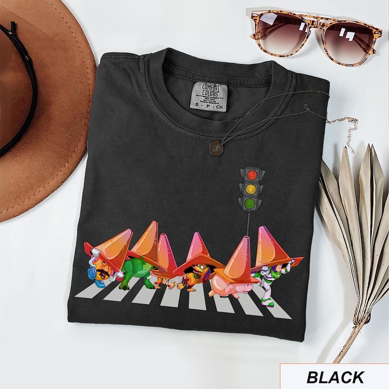 May include: Black t-shirt featuring a whimsical design of cartoon characters wearing traffic cones and crossing a crosswalk, with a traffic light above. The shirt includes the text "BLACK" at the bottom.