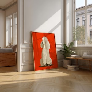 May include: A framed art print showing a white poodle illustration on a red background. The poodle is in a seated pose. The artwork is displayed in a room with wooden flooring and natural light streaming through a window.