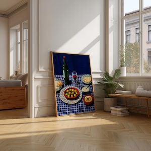 May include: A framed print depicting a still life with a table set for a meal. A green wine bottle and a glass of red wine sit on a blue checked tablecloth. Plates of olives, bread, and dips are also present. The artwork is displayed in a well-lit room.