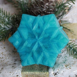 May include: A turquoise snowflake-shaped soap, with a detailed geometric design, rests on a decorative gift box. The soap is the focal point, with evergreen sprigs and a pinecone in the background, suggesting a winter or holiday theme.
