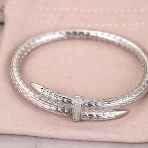 May include: A silver-colored bracelet with a textured, snake-like design. The bracelet features two pointed ends that overlap and are secured by a small band adorned with small, clear stones. The bracelet is resting on a light pink pouch.