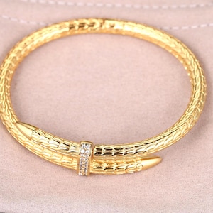 Puede incluir: Un brazalete dorado con un diseño texturizado en forma de serpiente. El brazalete presenta un cierre adornado con pequeñas piedras preciosas transparentes. Descansa sobre una tela beige claro.