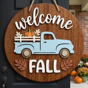 May include: A round wooden door sign with a dark brown wood grain finish. The sign features the white text "welcome" at the top and "FALL" at the bottom. A light blue truck with a white fence bed carries an orange pumpkin and beige leaves.