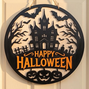 Peut inclure: Une décoration murale circulaire en bois avec un motif noir découpé au laser représentant une maison hantée, des arbres nus, des chauves-souris et des tombes sur un fond en bois clair. Le texte "HAPPY HALLOWEEN" est orange avec trois citrouilles.