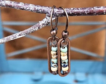 May include: A pair of handcrafted earrings with a copper-colored metal frame. Each earring features a vertical row of colorful beads in shades of blue, green, cream, and brown. The earrings are suspended from a simple hook.