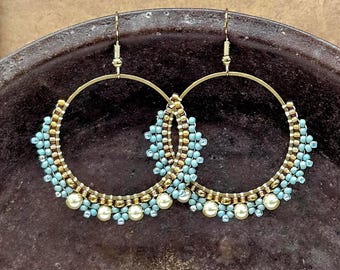 May include: A pair of gold-tone hoop earrings. The hoops are adorned with light blue and gold beads, and white pearl-like beads. The earrings are displayed on a dark brown surface.